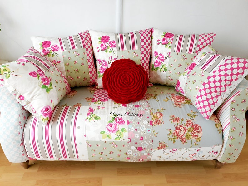 May include: A white and pink patchwork sofa with a red rose-shaped throw pillow. The sofa has a floral pattern and is decorated with several pillows.