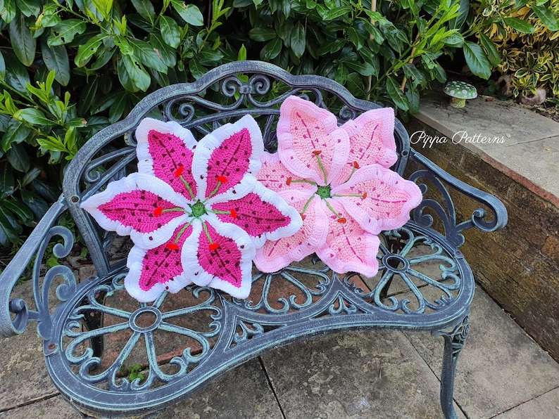 May include: Two crocheted lily flower cushions in pink and white on a black metal bench. The cushions are handmade and feature a detailed design.