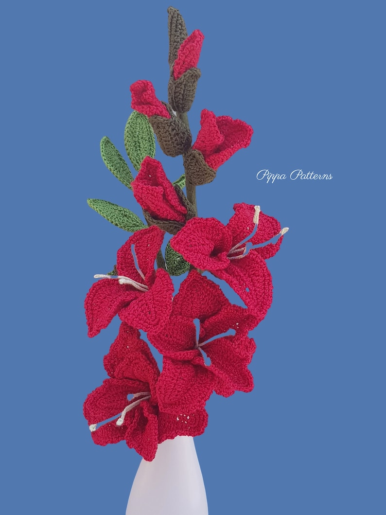 May include: A bouquet of crocheted red gladiolus flowers in a white vase. The flowers are arranged in a vase on a blue background. The text "Pippa Patterns" is visible in the upper right corner of the image.