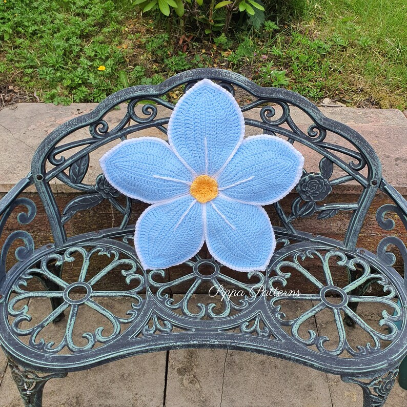 May include: A light blue crocheted flower cushion with a yellow center, sitting on a black metal bench.