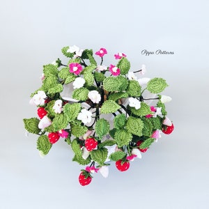 May include: A crocheted strawberry plant with pink and white flowers in a white pot. The plant is hanging from the pot and has green leaves. The text "Pippa Patterns" is visible in the upper right corner of the image.