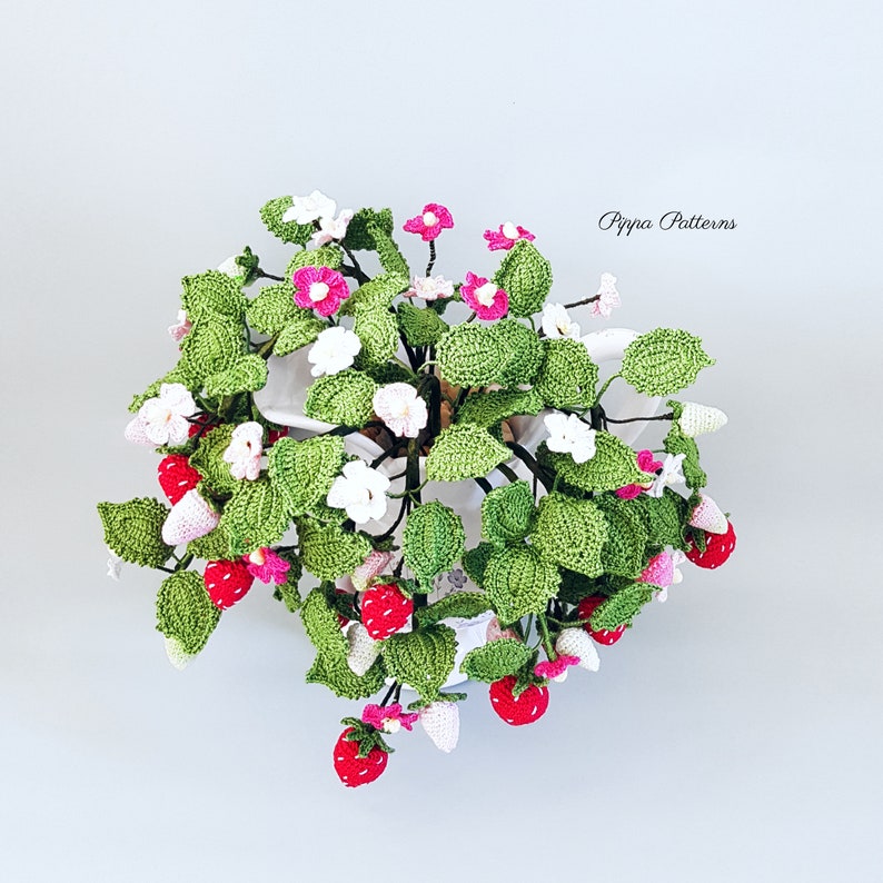 May include: A crocheted strawberry plant with pink and white flowers in a white pot. The plant is hanging from the pot and has green leaves. The text "Pippa Patterns" is visible in the upper right corner of the image.