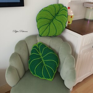 May include: Two green crocheted leaf-shaped pillows on a light green chair. The pillows have a raised texture and a dark green center.