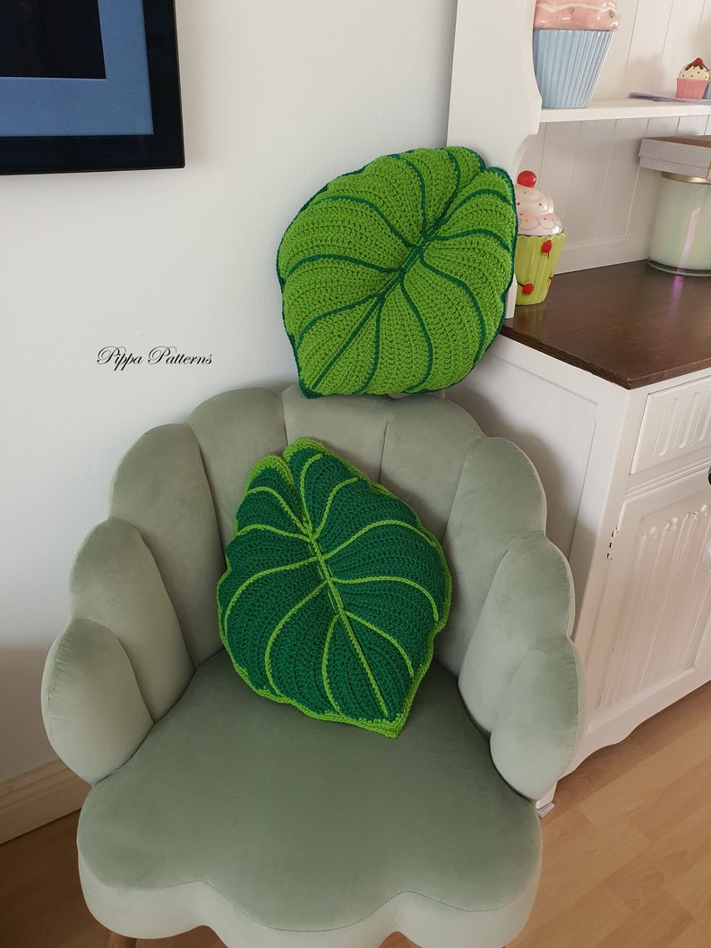 May include: Two green crocheted leaf-shaped pillows on a light green chair. The pillows have a raised texture and a dark green center.