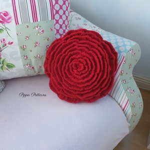 May include: A red crocheted rose cushion on a white couch with a floral patterned cushion behind it. The text "Pippa Patterns" is visible in the bottom left corner of the image.