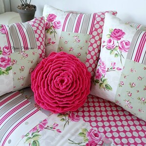 May include: A pink crocheted flower pillow on a bed with floral patterned pillows and a striped throw blanket. The pillows have a patchwork design with pink, white, and green floral patterns.