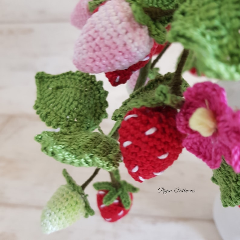 May include: A close-up of a crocheted strawberry plant with red, pink, and green strawberries and leaves. The plant is in a white pot and the text "Pippa Patterns" is visible in the bottom right corner.