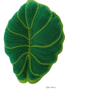 May include: A green crocheted leaf pillow with a light green outline. The pillow is shaped like a large leaf with a pointed tip and a rounded base.