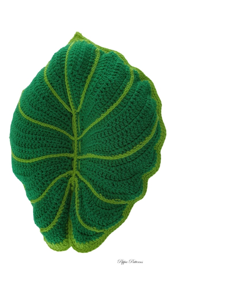 May include: A green crocheted leaf pillow with a light green outline. The pillow is shaped like a large leaf with a pointed tip and a rounded base.