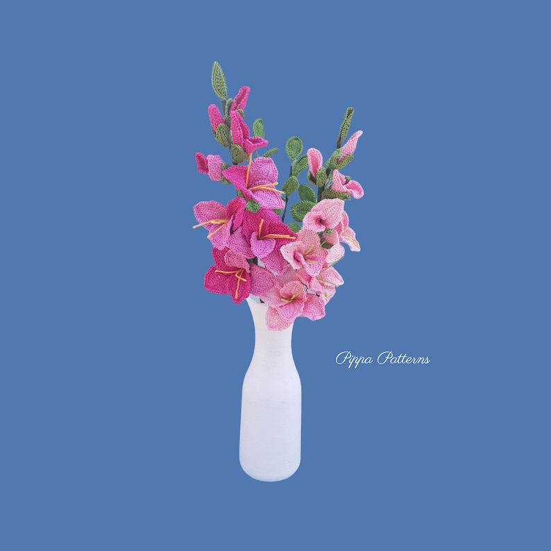 May include: A bouquet of pink and white crocheted gladiolus flowers in a white vase. The flowers are arranged in a vase with a simple design. The background is a solid blue color. The text "Pippa Patterns" is visible at the bottom of the image.