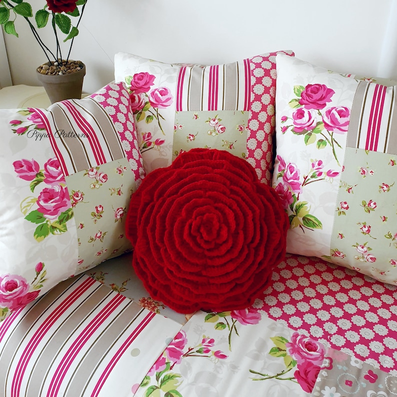 May include: A red, crocheted rose-shaped pillow sits on a patchwork couch with floral patterns in shades of pink, green, and white. The couch is made of a variety of fabrics, including stripes and polka dots.