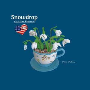 Può includere: Un arrangiamento di fiori di bucaneve all'uncinetto in una tazza da tè e un piattino blu. I fiori sono bianchi con steli e foglie verdi. La tazza da tè ha un motivo floreale e il piattino è di un blu pieno. Il testo "Snowdrop Crochet Pattern" e "PDF" è visibile nell'immagine.