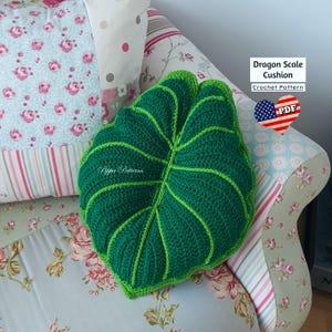 May include: Green crocheted leaf-shaped cushion with a textured surface. The cushion is on a floral patterned sofa. The text "Dragon Scale Cushion Crochet Pattern" is visible in the image.