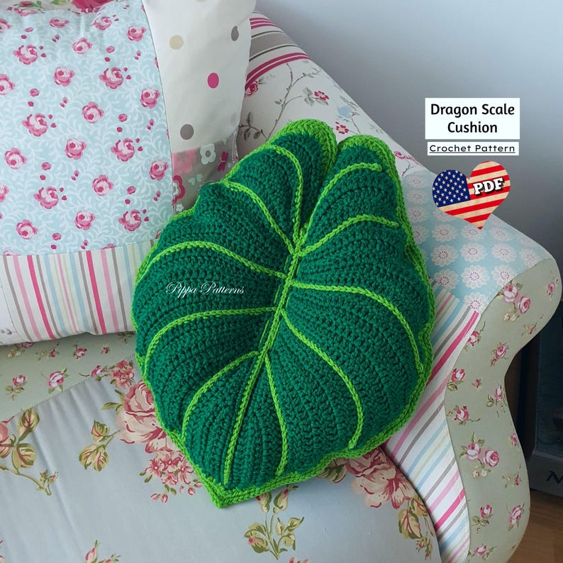 May include: Green crocheted leaf-shaped cushion with a textured surface. The cushion is on a floral patterned couch. The text "Dragon Scale Cushion Crochet Pattern" is visible in the image.