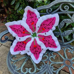 May include: A crocheted lily flower cushion in white and pink with green stems and red accents. The cushion is sitting on a metal bench with a floral design.