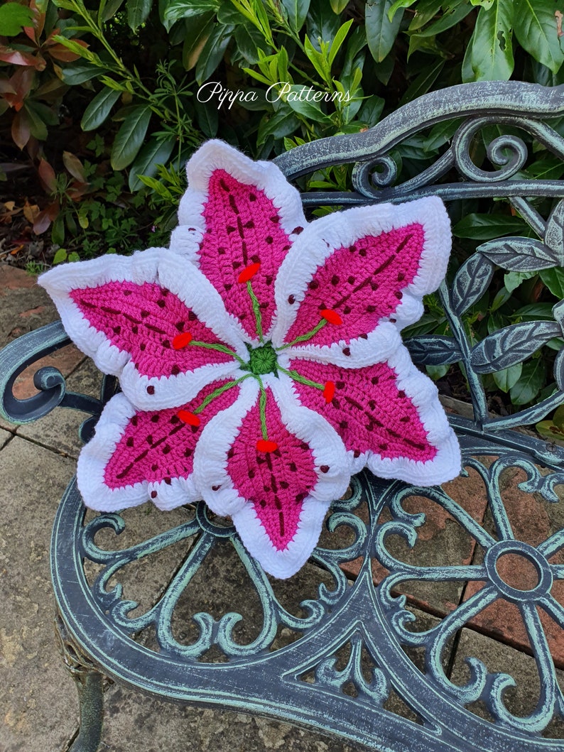 May include: A crocheted lily flower cushion in white and pink with green stems and red accents. The cushion is sitting on a metal bench with a floral design.