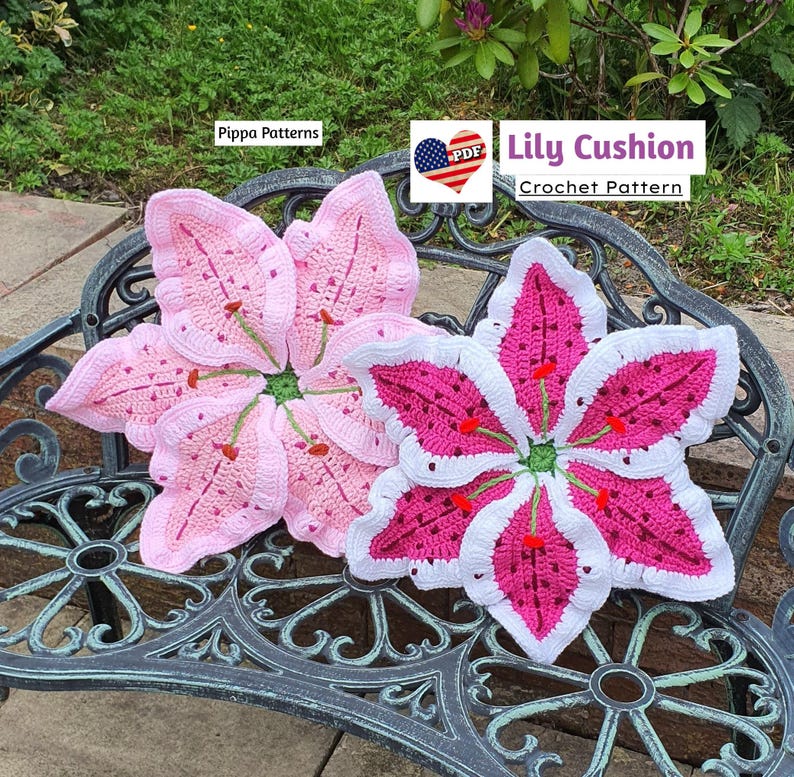 May include: Two crocheted lily flower cushions, one pink and one white and pink, on a black metal bench. The pink cushion has a green center and red accents. The white and pink cushion has a green center and red accents. The text "Lily Cushion Crochet Pattern" is visible in the image.