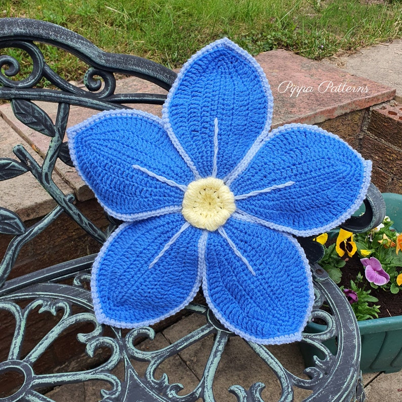 May include: A crocheted blue flower with a yellow center, resting on a black metal bench. The flower is a forget-me-not design and has a white outline. The text "Pippa Patterns" is visible in the background.