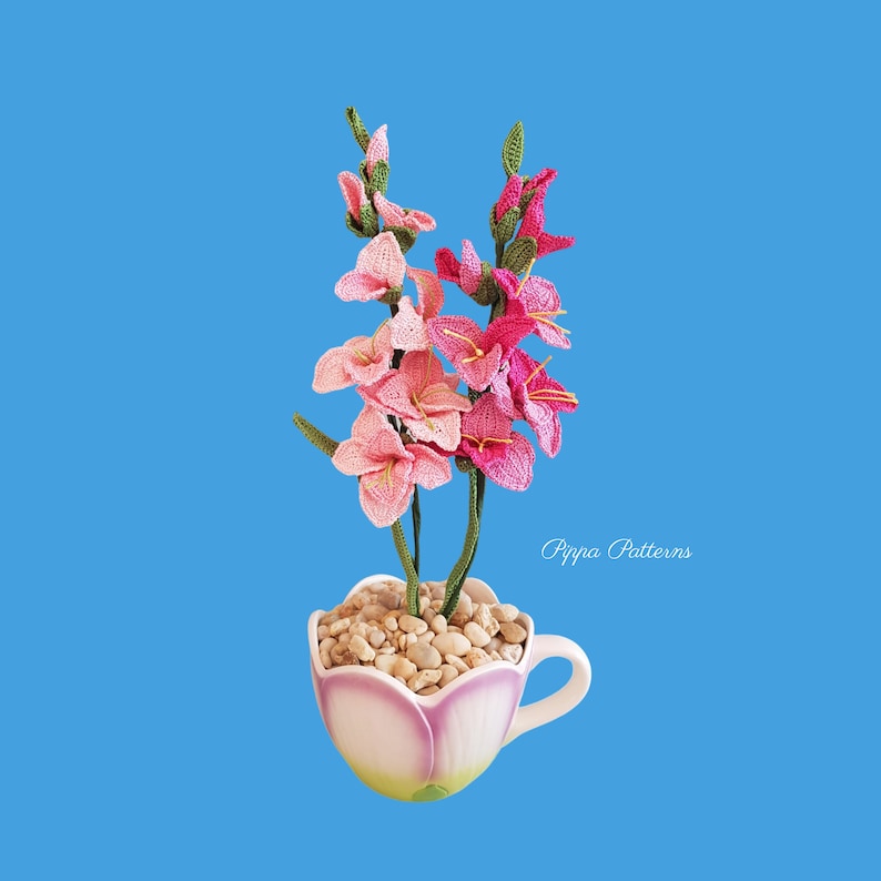 May include: A crocheted pink and white gladiolus flower arrangement in a white ceramic teacup with white stones. The teacup has a handle and a floral design. The text "Pippa Patterns" is visible in the image.