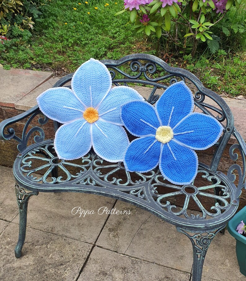 May include: Two crocheted blue flower cushions with yellow centers, sitting on a black metal garden bench. The bench is on a stone patio.