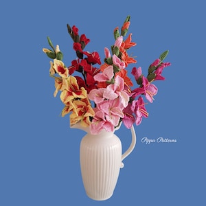 May include: A bouquet of crocheted gladiolus flowers in shades of pink, red, yellow, and orange arranged in a white ceramic pitcher. The flowers are arranged in a vase with a ribbed design. The text "Pippa Patterns" is visible in the bottom right corner of the image.