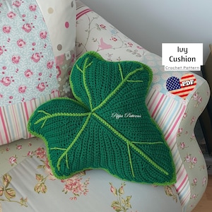 Może przedstawiać: Zielona, dziana poduszka w kształcie liścia bluszczu z jasnozieloną obwódką. Poduszka leży na kanapie z kwiatowym wzorem. Tekst "Ivy Cushion Crochet Pattern PDF" znajduje się w prawym górnym rogu obrazu.
