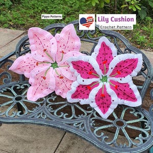 May include: Two crocheted lily flower cushions, one pink and one white with pink accents, on a metal bench. The pink cushion has a white border. The white cushion has a pink center and a white border. The text "Lily Cushion Crochet Pattern" is visible in the image.