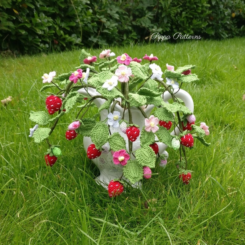 May include: A crocheted strawberry plant with pink and white flowers in a white pitcher. The plant is sitting on a bed of green grass. The text "Pippa Patterns" is visible in the upper right corner of the image.