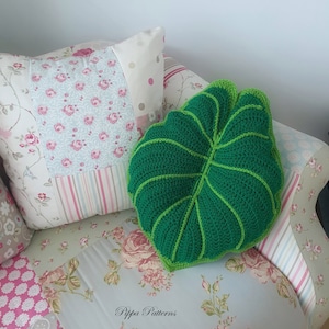 May include: A green crocheted leaf-shaped pillow on a floral patterned couch. The pillow has a light green outline and is sitting on a couch with a floral pattern.