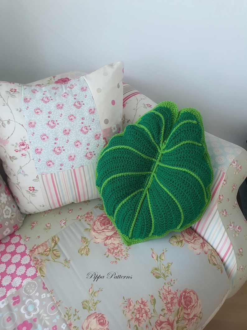 May include: A green crocheted leaf-shaped pillow on a floral patterned couch. The pillow has a light green outline and is sitting on a couch with a floral pattern.