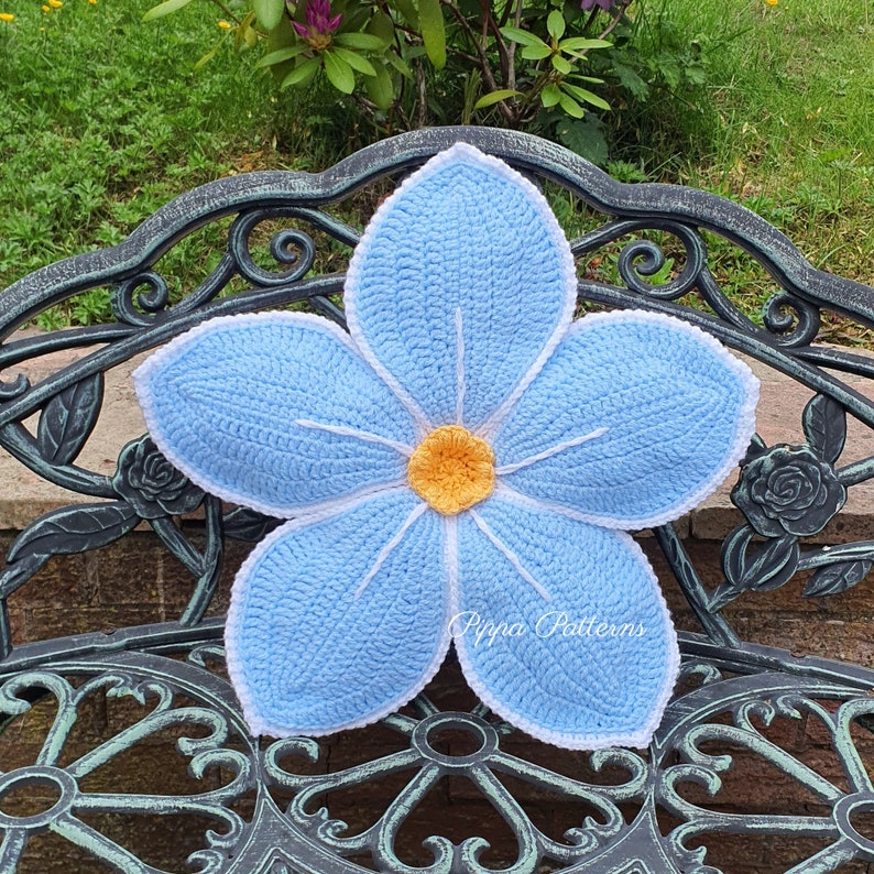 May include: A crocheted blue flower cushion with a yellow center, resting on a black metal garden bench.
