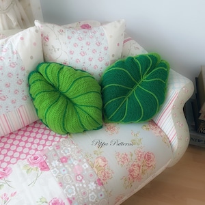 May include: Two green crocheted leaf-shaped pillows on a floral patterned couch. The pillows are on a couch with a patchwork floral design. The text "Pippa Patterns" is visible on the couch.