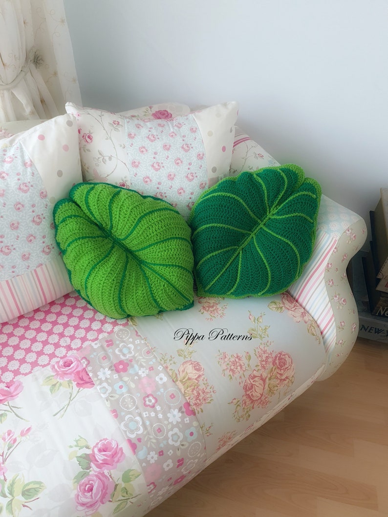 May include: Two green crocheted leaf-shaped pillows on a floral patterned couch. The pillows are on a couch with a patchwork floral design. The text "Pippa Patterns" is visible on the couch.