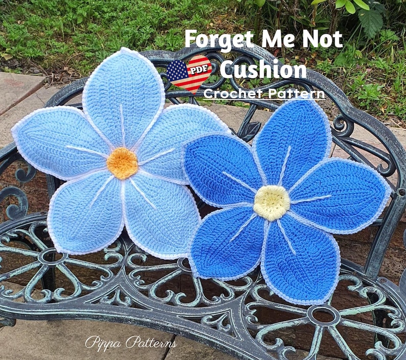 May include: Two crocheted forget-me-not flower cushions in blue and light blue with white centers. The cushions are on a black metal bench. The text "Forget Me Not Cushion Crochet Pattern" is above the cushions.