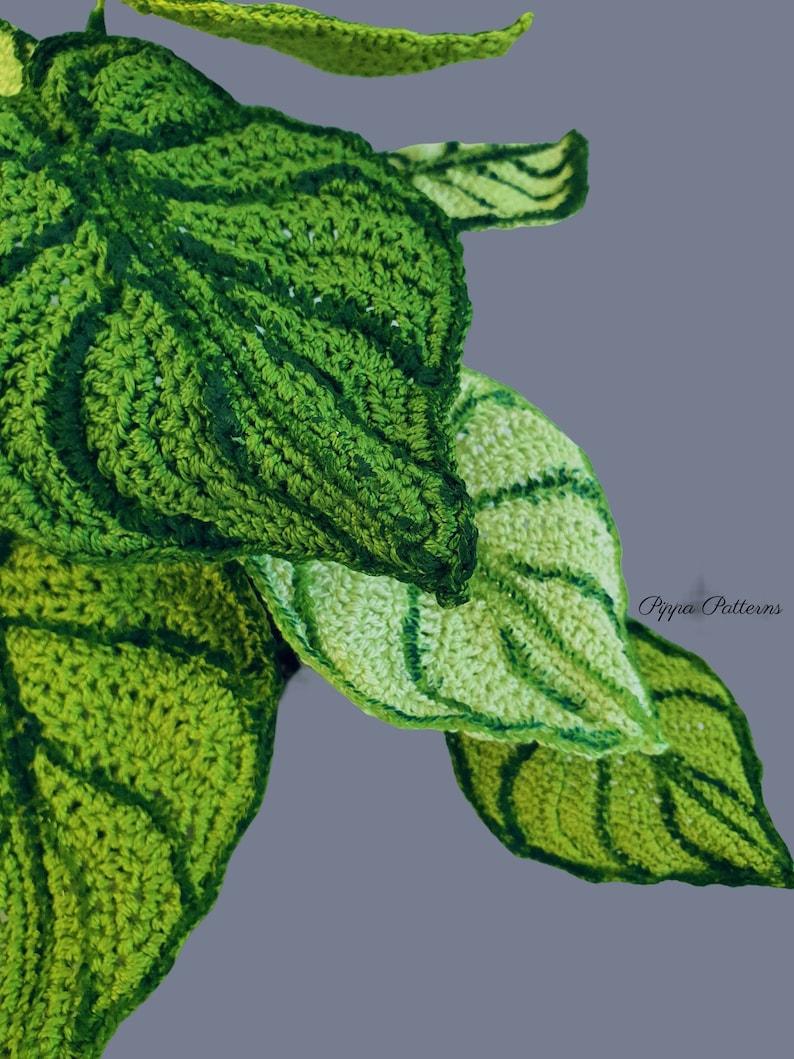 May include: Close-up of a crocheted green leaf plant with a textured surface. The leaves are layered on top of each other and have a light green and dark green color variation. The text "Pippa Patterns" is visible in the bottom right corner of the image.