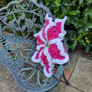 May include: A pink and white crocheted lily flower pillow on a black metal bench. The pillow is sitting on a patio with a green bush in the background. The text "Pippa Patterns" is visible in the upper right corner of the image.