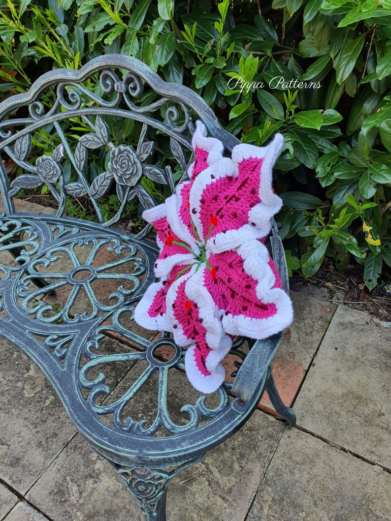 May include: A pink and white crocheted lily flower pillow on a black metal bench. The pillow is sitting on a patio with a green bush in the background. The text "Pippa Patterns" is visible in the upper right corner of the image.