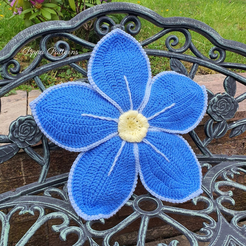May include: A crocheted blue flower cushion with a white center. The cushion is sitting on a black metal bench.