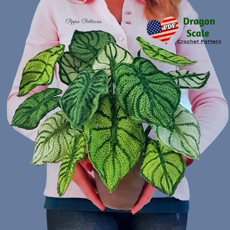 May include: A crocheted plant with green and white leaves in a brown pot. The plant is being held by a person wearing a pink shirt. The text "Dragon Scale Crochet Pattern" is in the top right corner of the image.