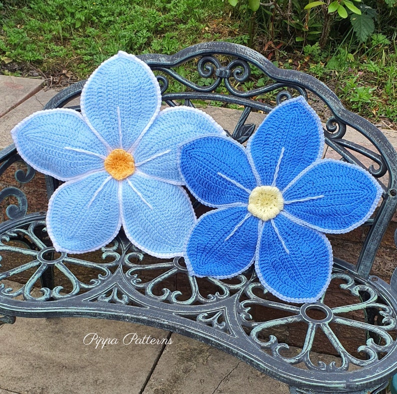 May include: Two crocheted blue flower cushions with white edging, sitting on a black metal bench. The flowers have yellow centers and are arranged in a symmetrical pattern.
