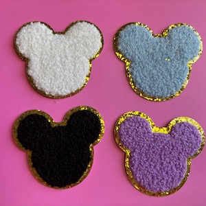 May include: Four embroidered mouse head patches. The patches are white, light blue, black, and purple, each with a gold glitter border. They are displayed on a pink background.