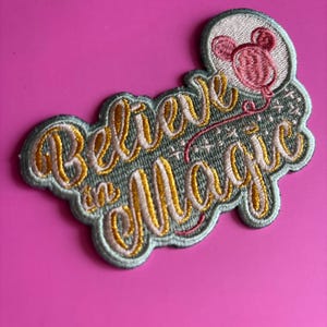 May include: Embroidered patch with the words "Believe in Magic" in a cursive font, outlined in gold thread. A pink mouse head with a balloon is in the upper right corner. The patch has a light green border and is set against a bright pink background.