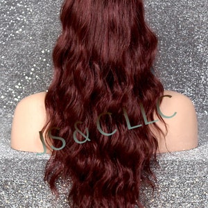 May include: A long, wavy wig in a deep burgundy color. The wig is styled with loose waves and has a natural-looking hairline. The hair is thick and appears to be made of synthetic fibers. The wig is displayed on a mannequin head against a sparkly silver background.