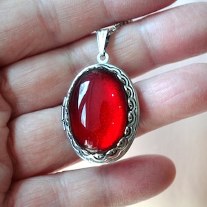 May include: A silver locket necklace with a red glass oval pendant. The locket has a detailed silver border.