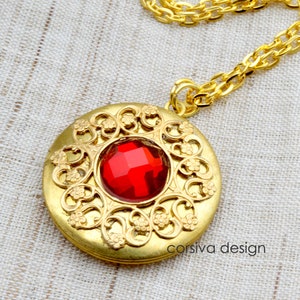 May include: A gold locket necklace with a red gemstone in the center. The locket is surrounded by a decorative filigree design. The necklace is on a gold chain.