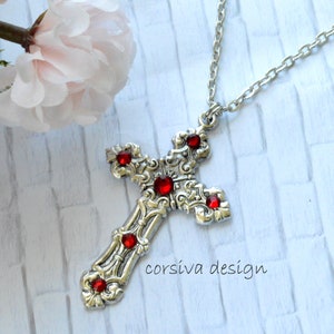 Puede incluir: Collar de cruz de plata con detalles de cristal rojo. La cruz tiene un diseño detallado y está en una cadena de plata.