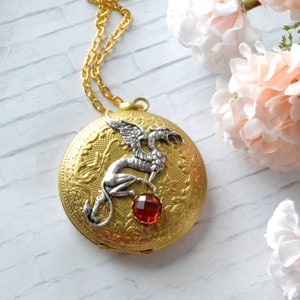 Pode incluir: Um colar com um medalhão de ouro adornado com um dragão prateado e uma pedra preciosa vermelha. O medalhão é gravado com um desenho floral.