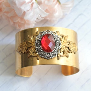 May include: A gold metal cuff bracelet with an ornate design and a large red gemstone in the center.