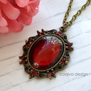 May include: A red glass cabochon pendant necklace with a gold-toned ornate frame and red rhinestone accents. The necklace is on a gold-toned chain.
