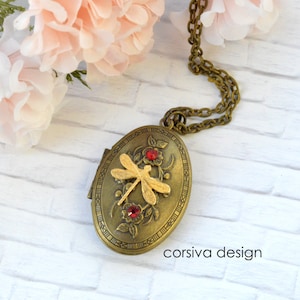 Puede incluir: Un collar con un medallón ovalado en tono dorado, adornado con un diseño de libélula y acentos de pedrería roja. El medallón está abierto y la cadena es visible. El medallón está sobre una superficie blanca.